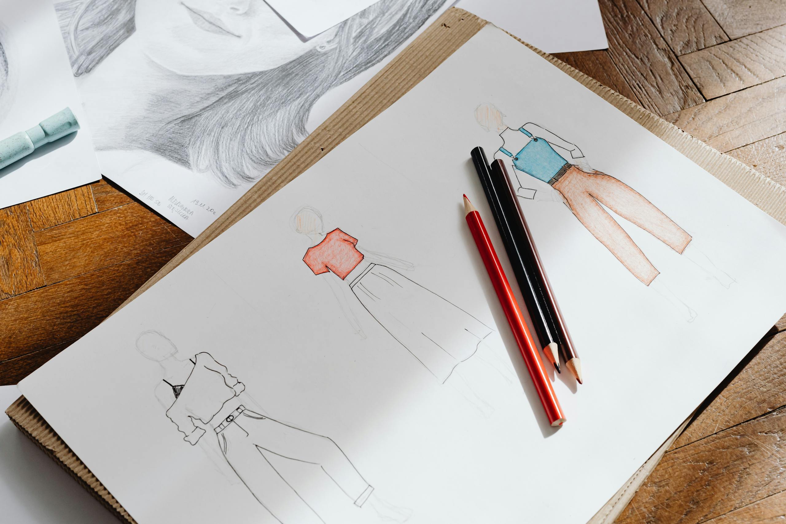 Artistic fashion design sketches on paper accompanied by colored pencils on a wooden surface.