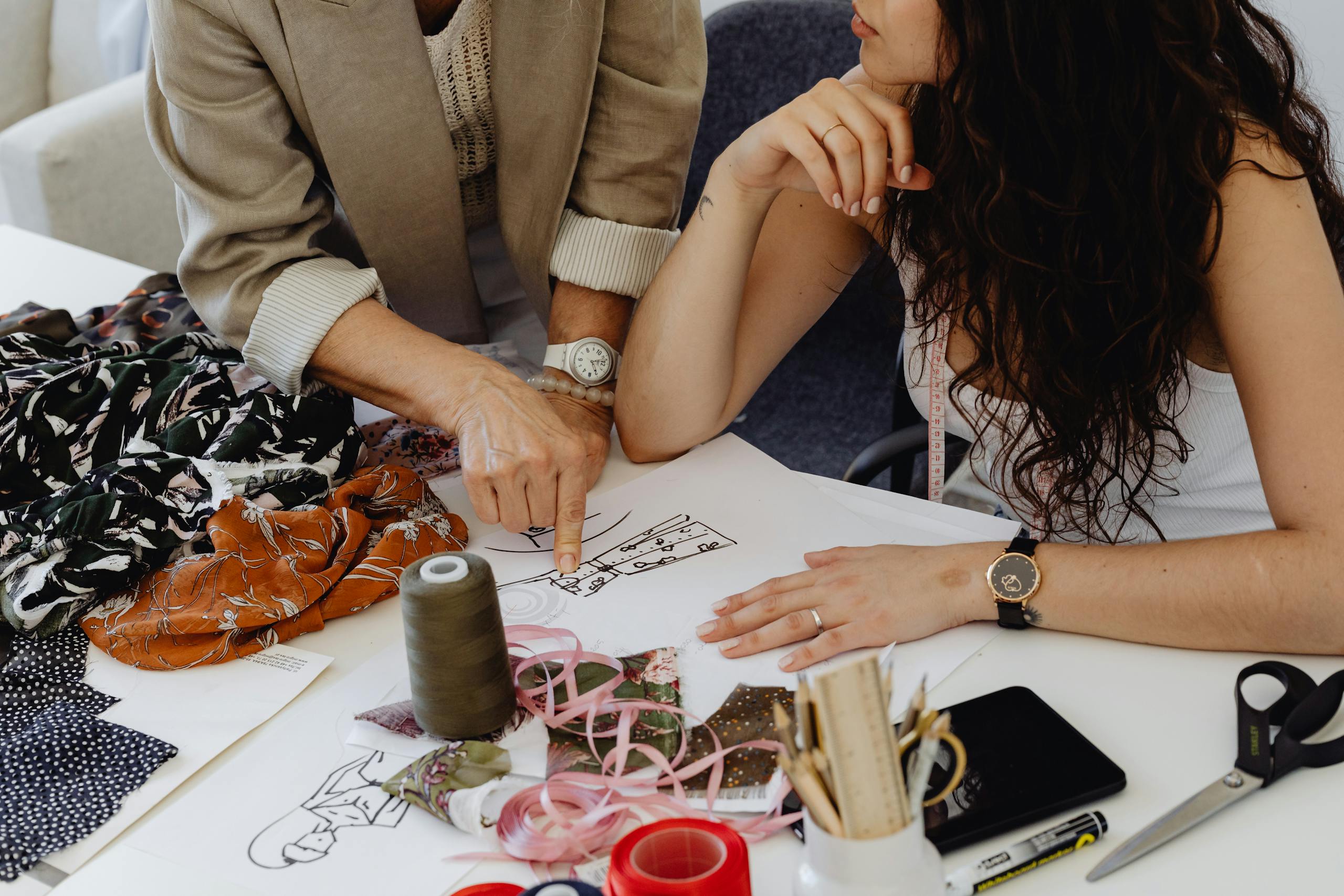 Two women collaborate on fashion sketches, fabrics, and tools, showcasing creative design process.